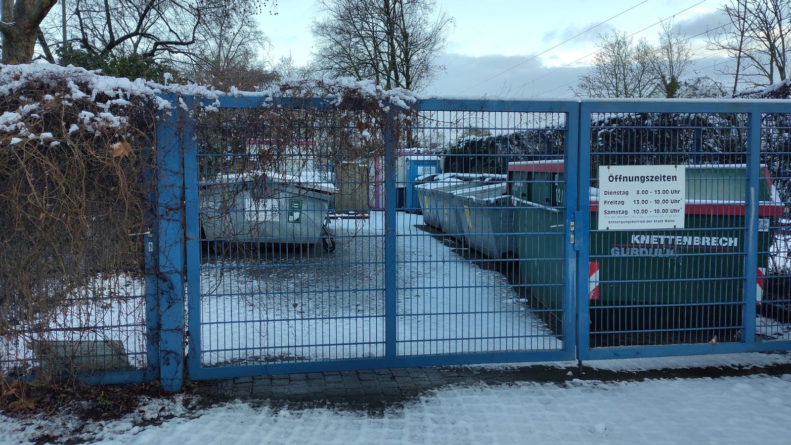 Eingangstor eines Wertstoffhofs mit Öffnungszeiten-Schild und Containern im Hintergrund