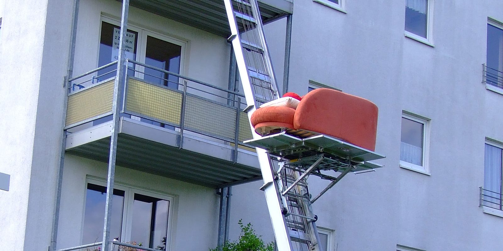 Sofa auf der Plattform eines Möbellifts, der schräg an einem Wohnhaus mit Balkonen lehnt